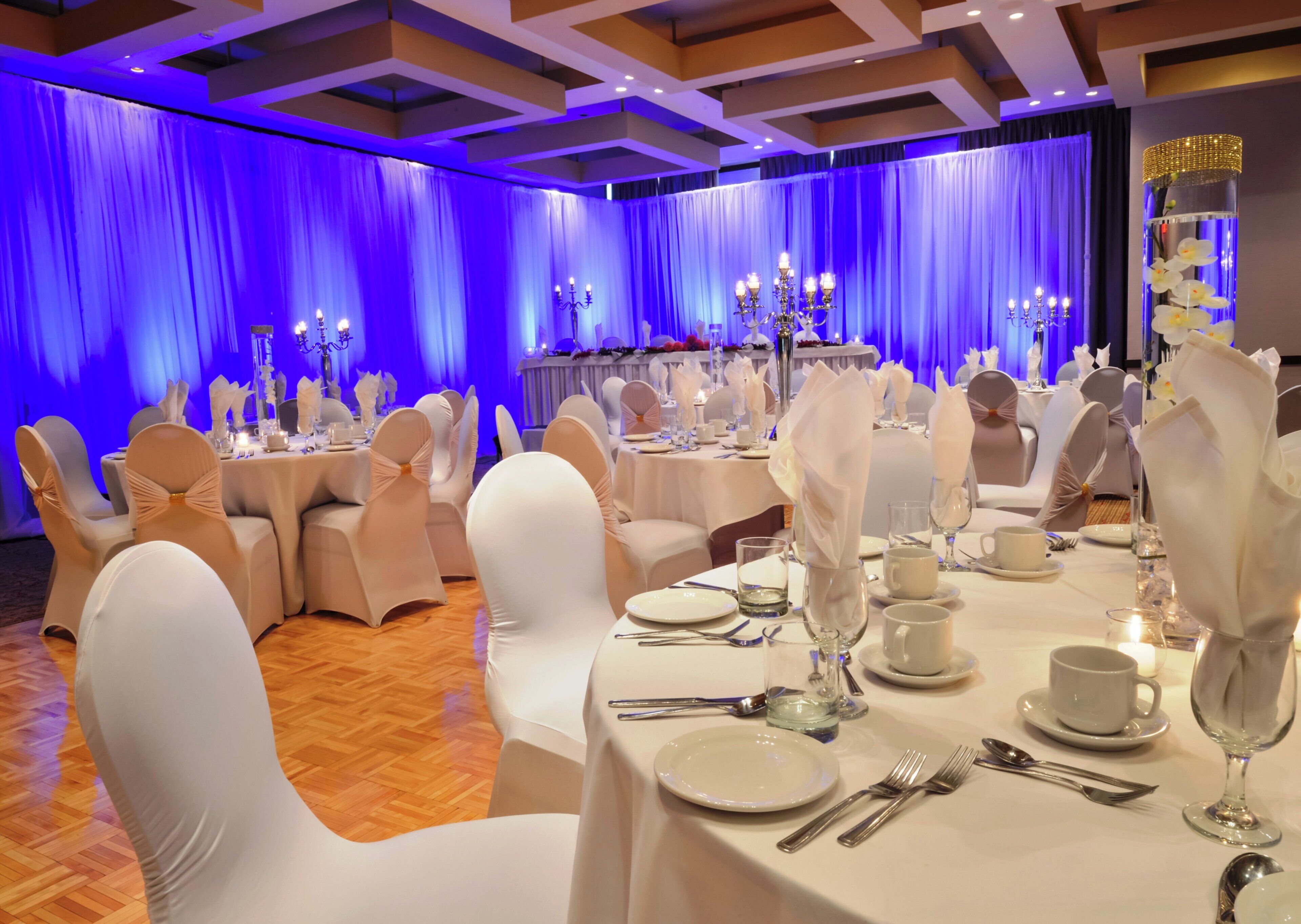 Un salón de banquetes con mesas redondas dispuestas para un evento formal, con manteles blancos, sillas con lazos y candelabros.