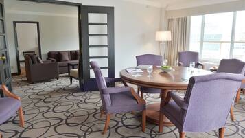 A hotel room with a round table, six chairs, a sofa, a coffee table, and a large window with curtains.