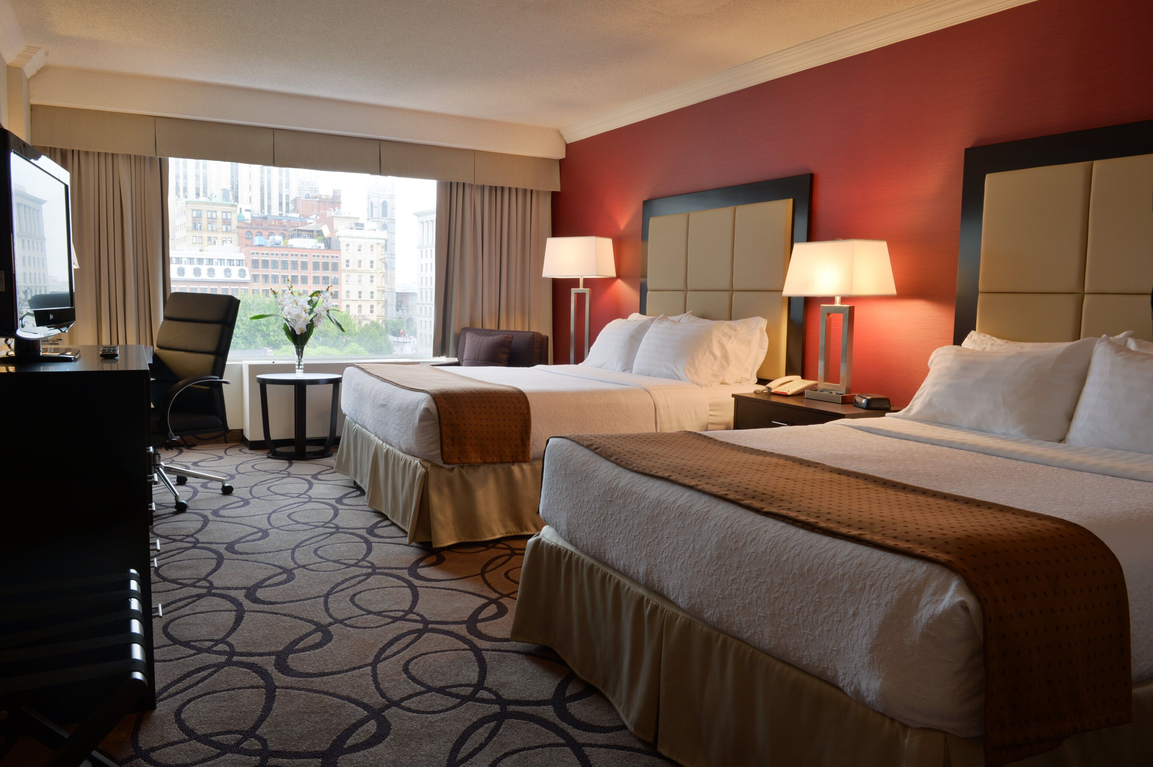 A hotel room with two beds, a desk, a chair, a lamp, and a view of the city.