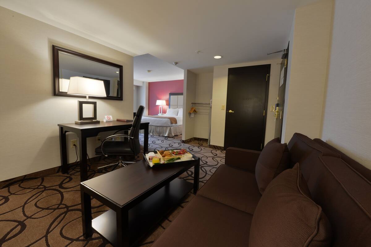 A hotel room with a brown sofa, a desk with a lamp, a bed with a white headboard, and a dining table with a tray of food.