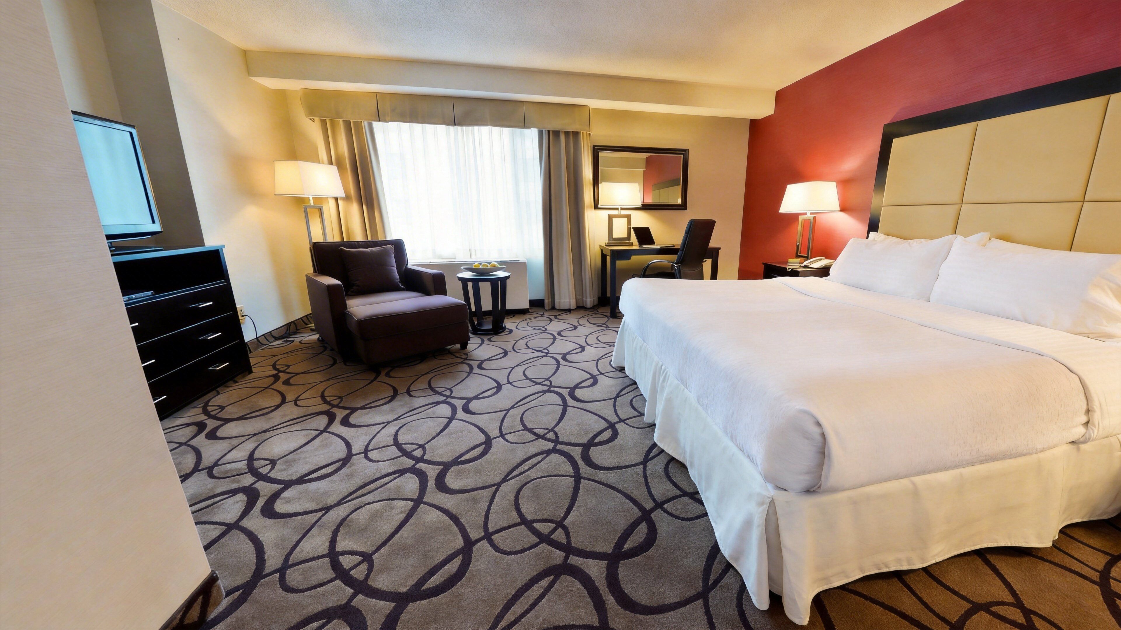 A hotel room with a bed, a desk, a chair, a TV, and a dresser.