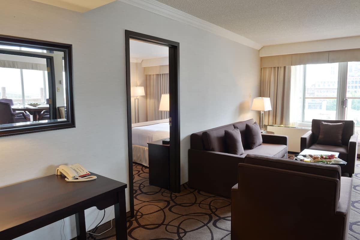 A hotel room with a bed, a sitting area with a sofa and armchairs, a desk with a telephone, and a large window with curtains.
