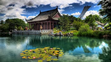 A traditional pavilion by a calm pond with lily pads, surrounded by lush greenery and a clear sky.