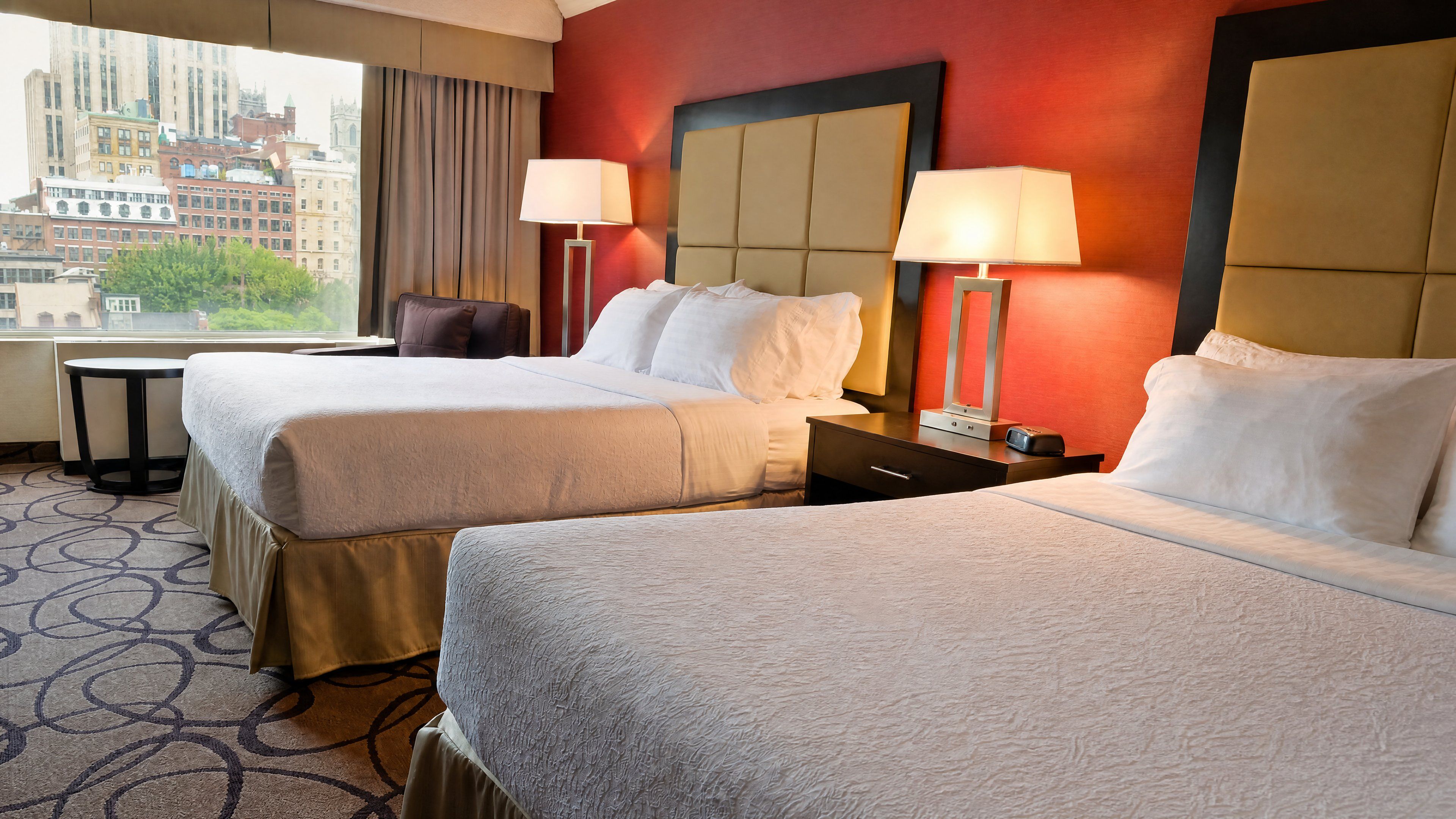 A hotel room with two beds, a desk, a chair, and a view of a cityscape.