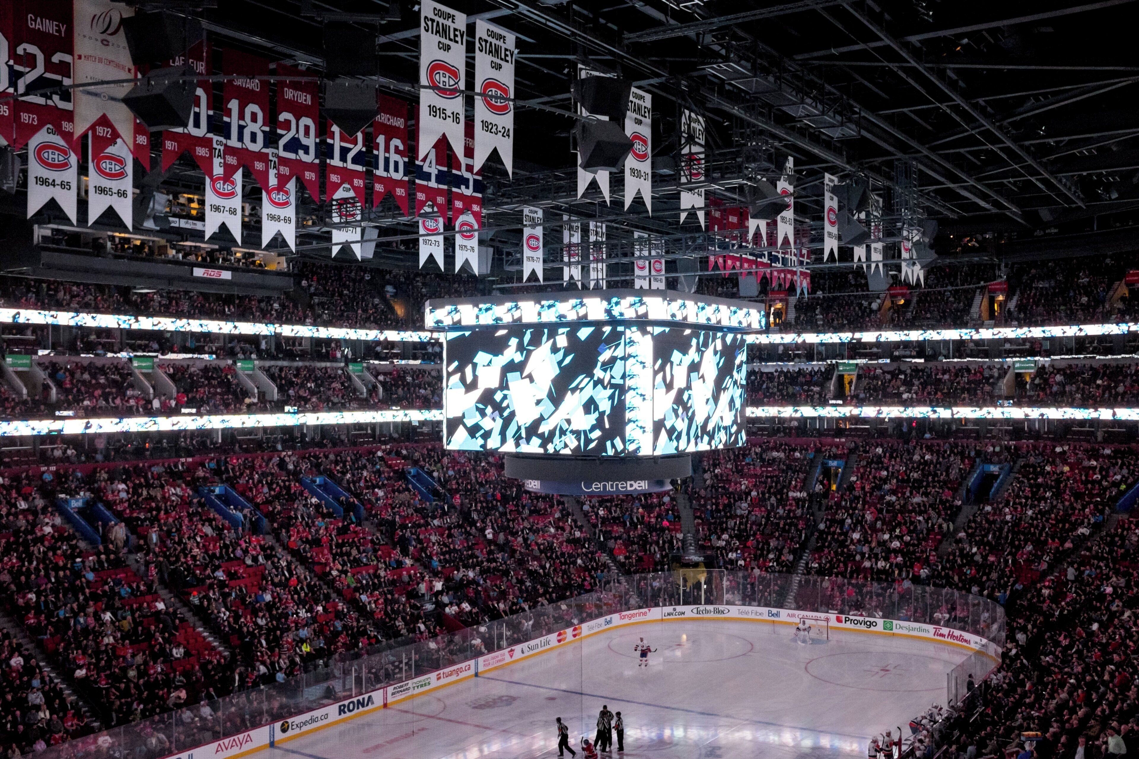 A large indoor arena with multiple levels of seating, a central ice rink, and a large screen displaying graphics.