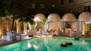 Zona de piscina interior con sillas y mesas blancas, rodeada de palmeras y áreas de descanso cubiertas.