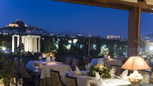 Un espacio de comedor al aire libre con mesas dispuestas para una comida, con vistas a una ciudad que cuenta con una colina destacada y arquitectura clásica.
