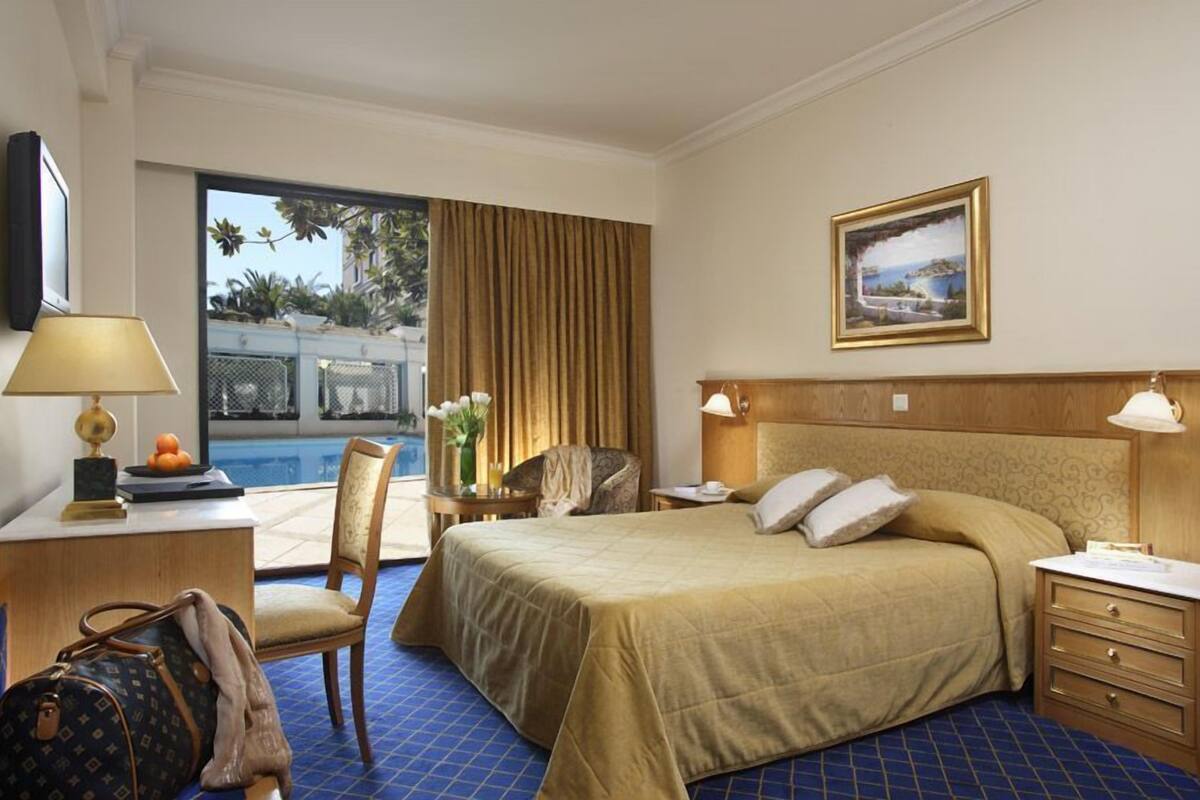 Una camera d'hotel con un letto, comodini, una scrivania con lampada, una sedia e vista su una piscina.