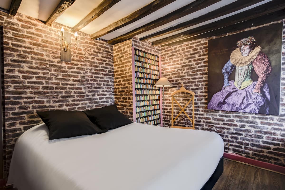Une chambre avec un mur en briques, une bibliothèque, un lit avec des oreillers noirs et un tableau représentant une figure historique.