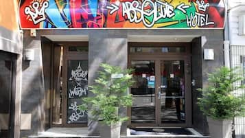 Ein Hotelzugang mit einem farbenfrohen Graffiti-Schild über der Tür, je einem Blumentopf auf beiden Seiten und einer Glastür, die das Innere reflektiert.