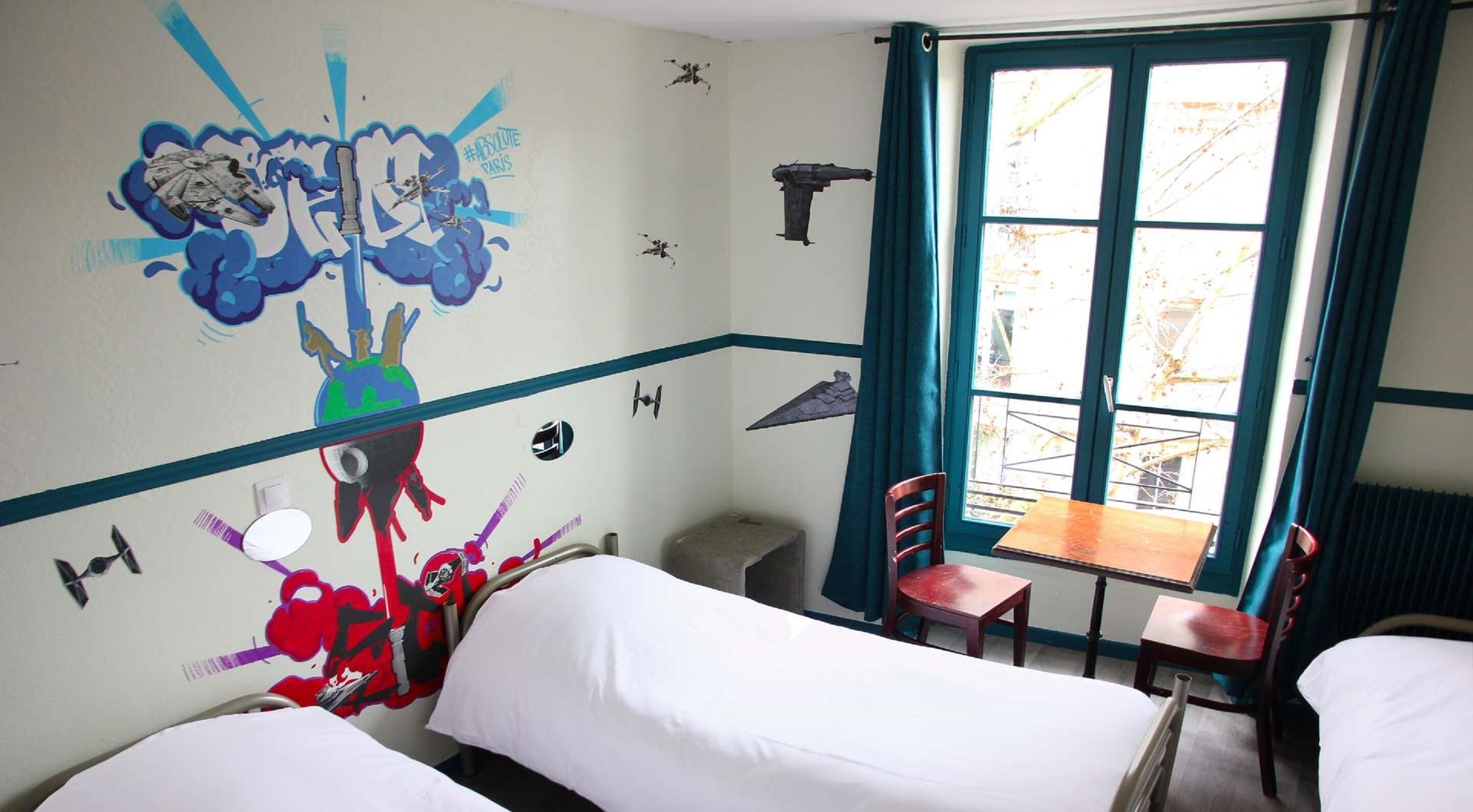 Habitación de hotel con dos camas, una ventana con vistas y un mural al estilo grafiti en la pared.