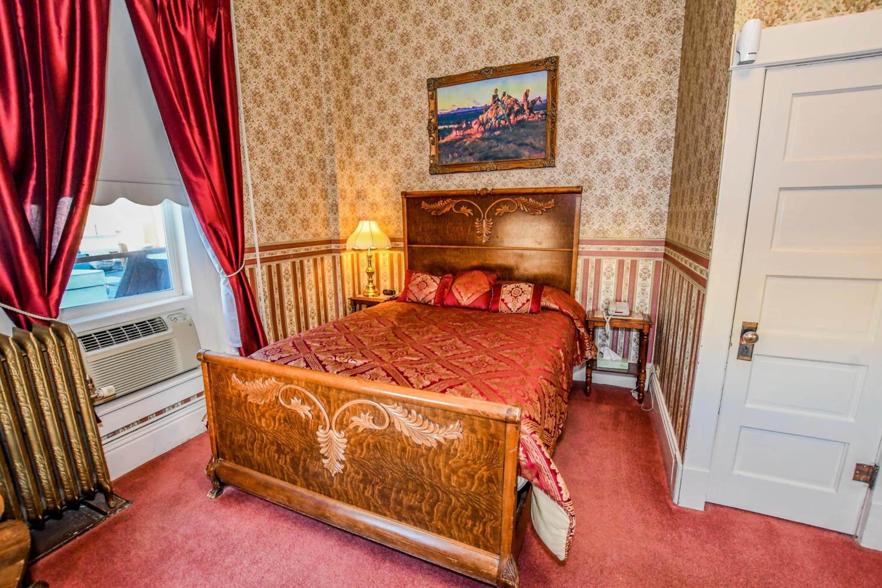 A bedroom with a wooden bed, red curtains, a radiator, a lamp, and a painting on the wall.