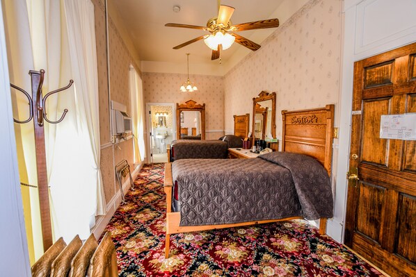 Quarto de hotel com cama, ventoinha no teto, porta de madeira e tapete estampado com flores.