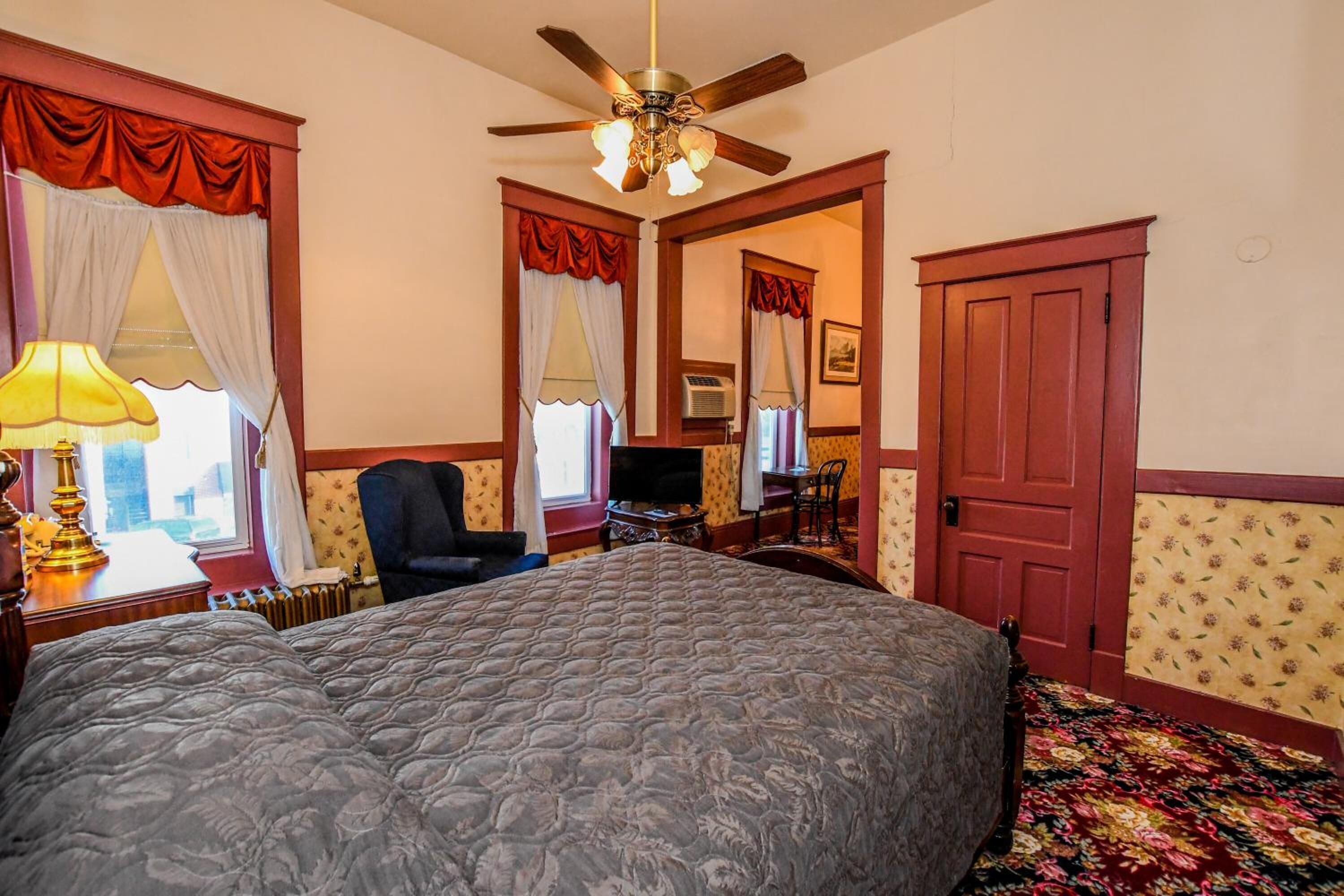 A bedroom with a large bed, a ceiling fan, a desk with a chair, a TV, and a door.