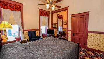 A bedroom with a large bed, a ceiling fan, a desk with a chair, a TV, and a door.