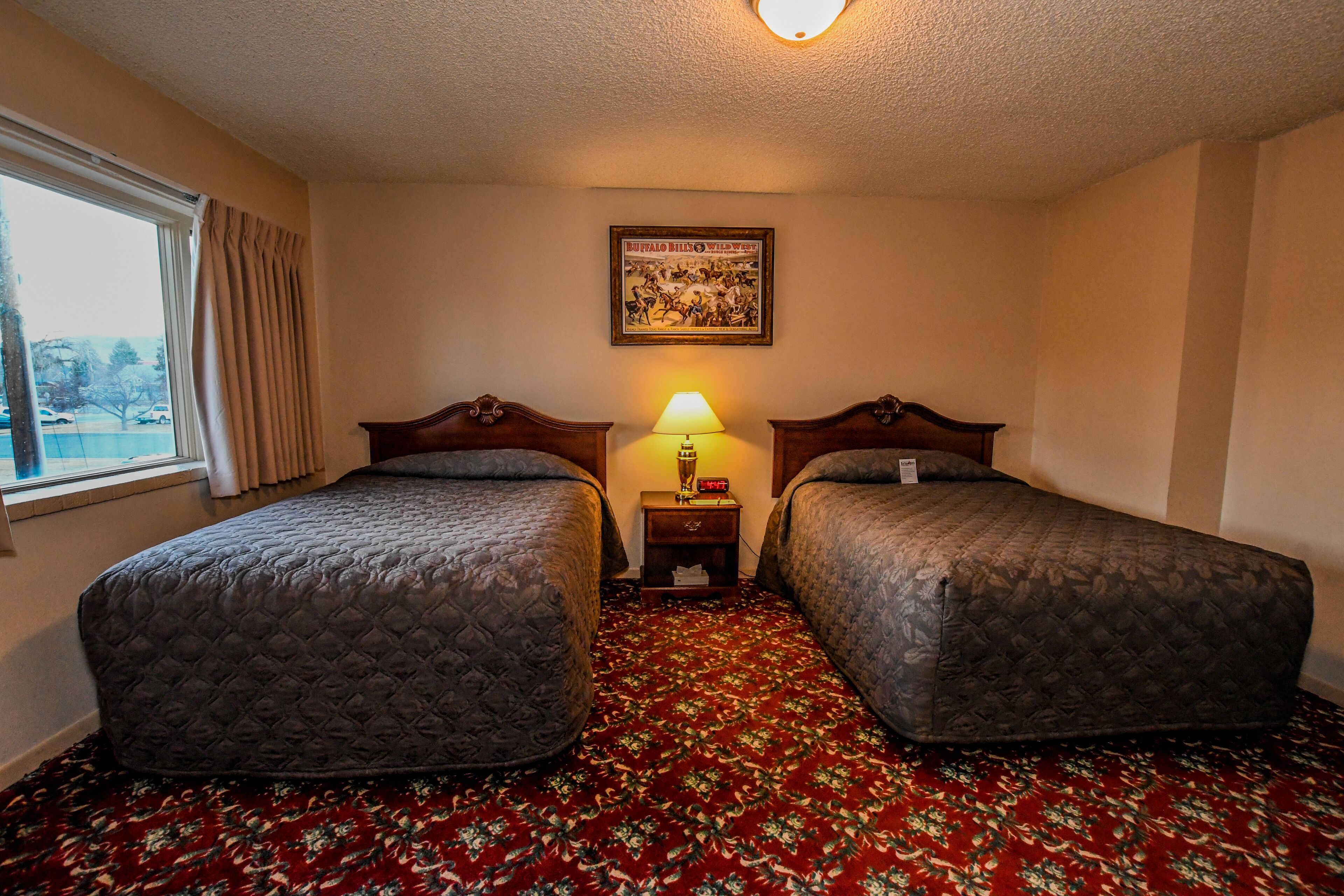 A hotel room with two beds, a floral carpet, a lamp, and a framed picture on the wall.