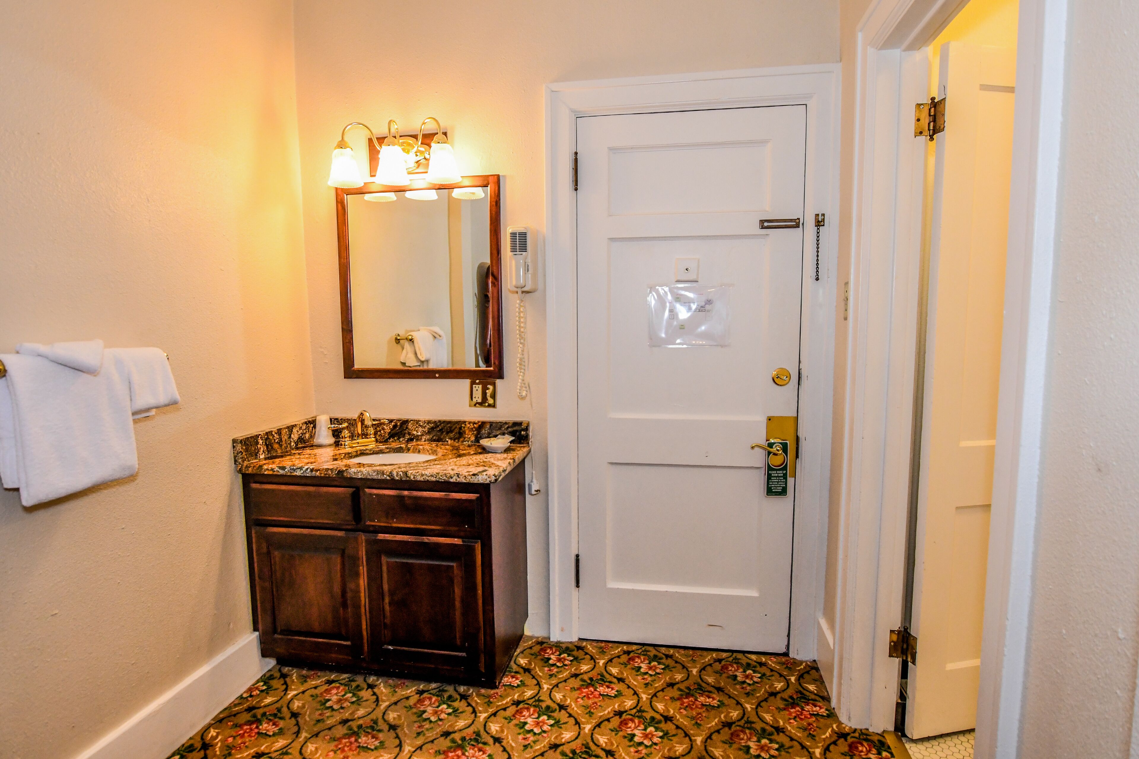 A bathroom with a wooden vanity, a mirror with lights, a white door, and a patterned floor.