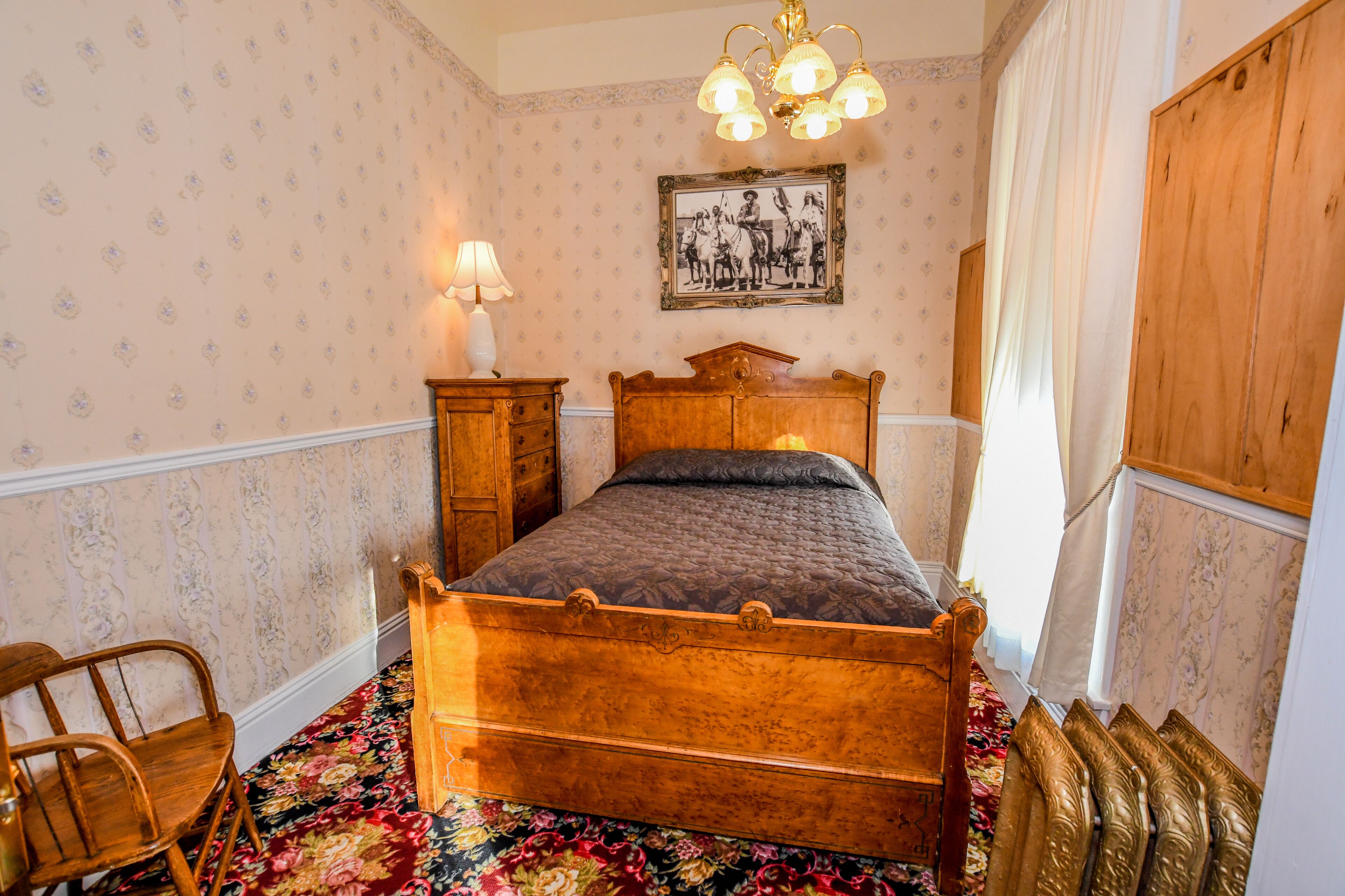 A bedroom with a wooden four-poster bed, a nightstand with a lamp, a floral carpet, and a framed picture on the wall.