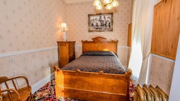 A bedroom with a wooden four-poster bed, a nightstand with a lamp, a floral carpet, and a framed picture on the wall.
