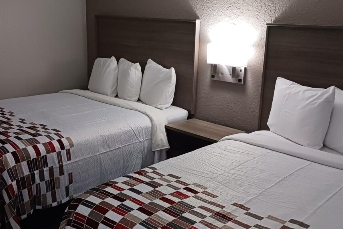 Quarto de hotel com duas camas, cada uma com colcha acolchoada, cabeceira de madeira e luminária fixada na parede.