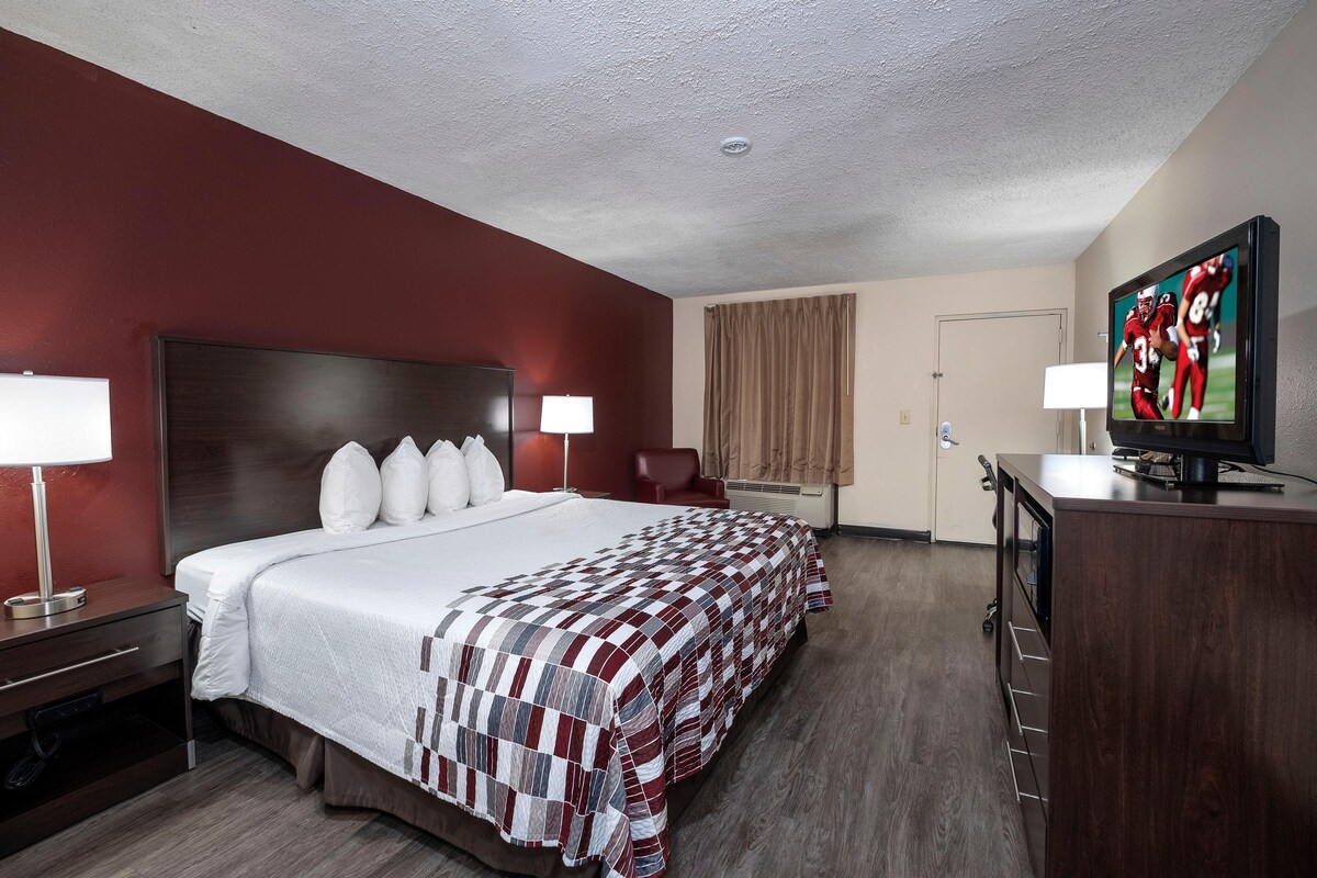 Quarto de hotel com uma cama grande, uma televisão em um suporte, uma escrivaninha com abajur e um guarda-roupa.