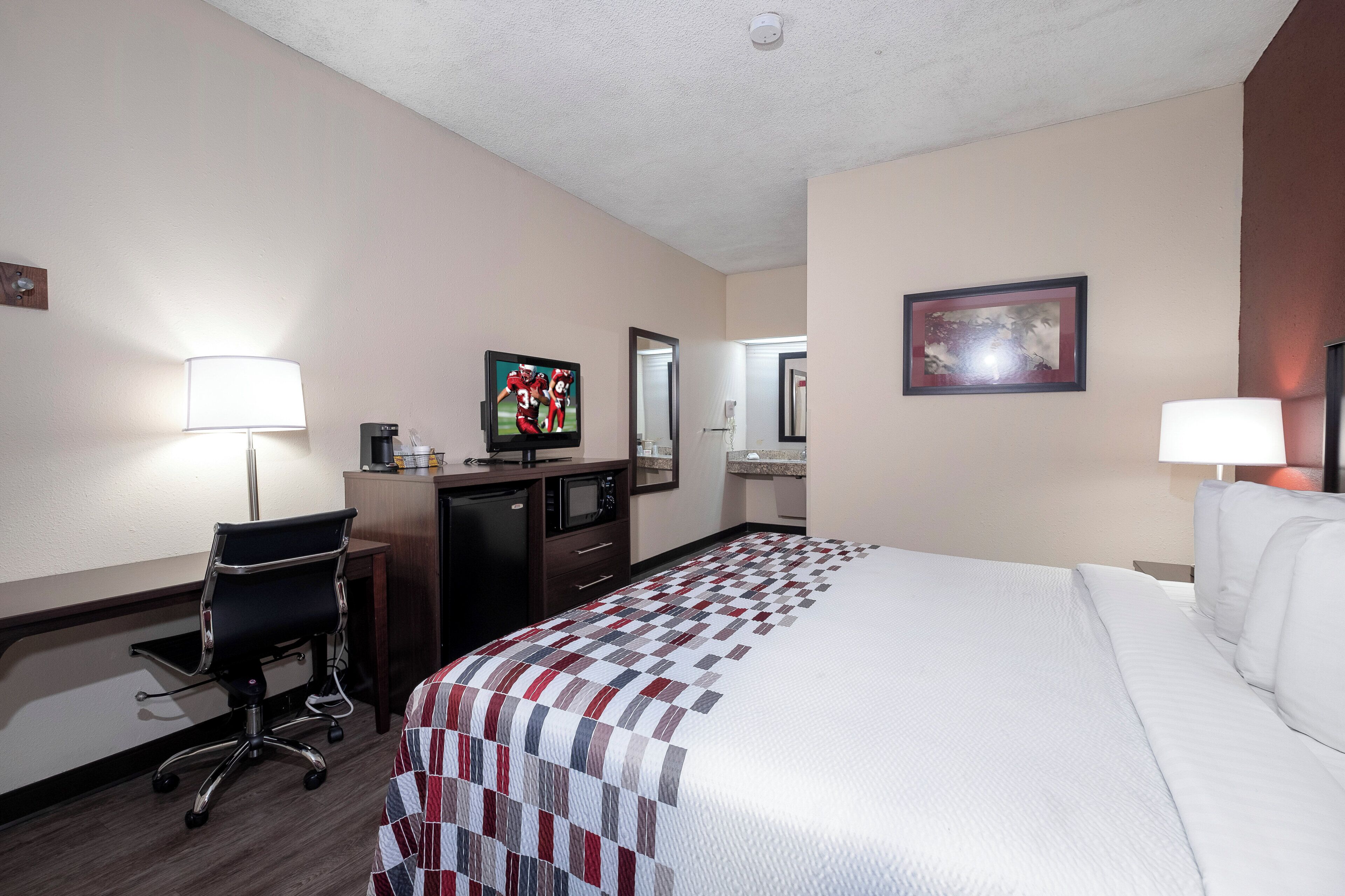 A hotel room with a bed, desk, chair, and a television on a stand.