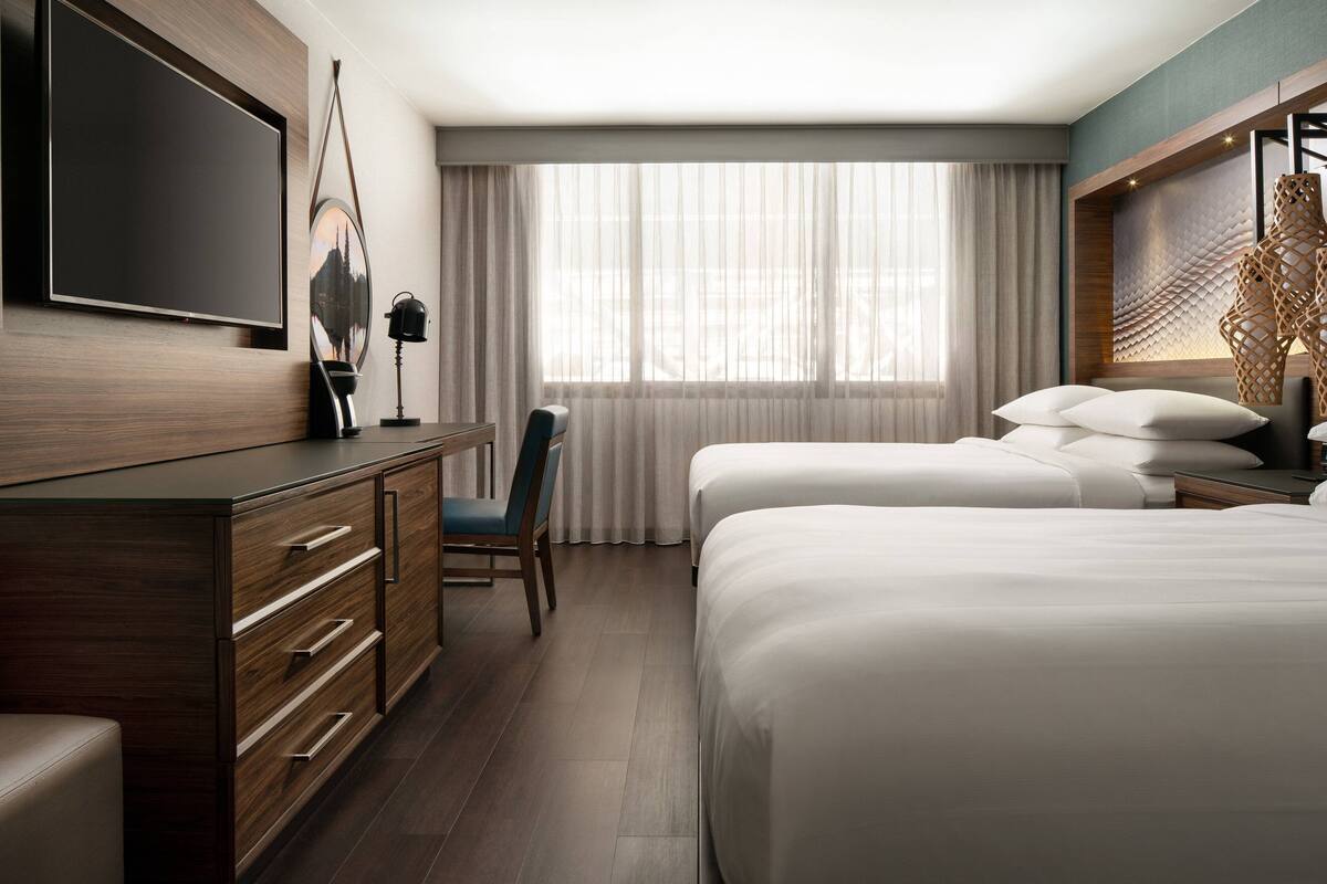 A hotel room with a large flat-screen TV mounted on the wall, a desk with drawers, a chair, two beds with white bedding, a window with sheer curtains, and a decorative wall panel.