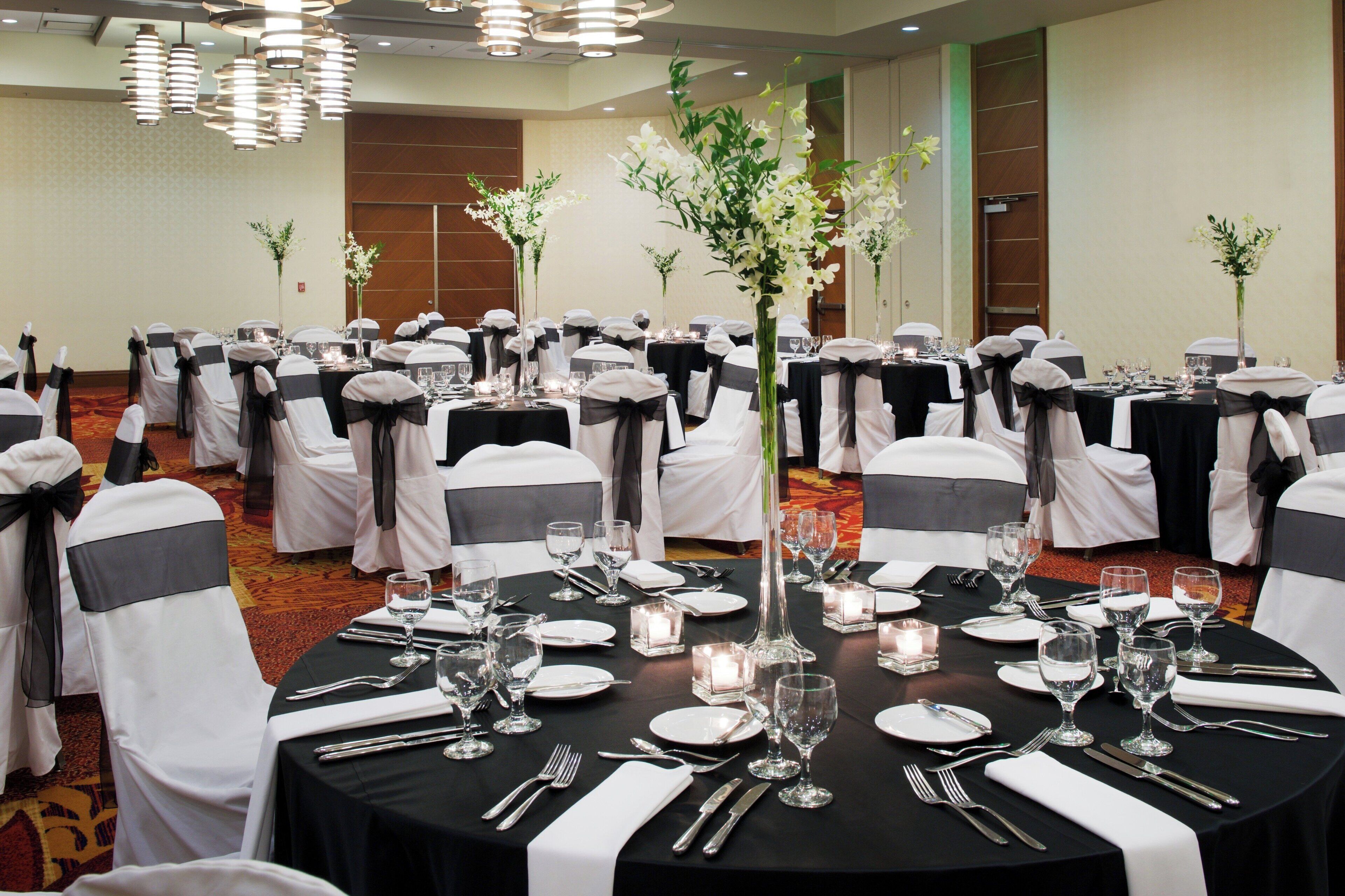 A banquet hall with round tables set for a formal event, featuring black tablecloths, white chairs with black sashes, and floral centerpieces.