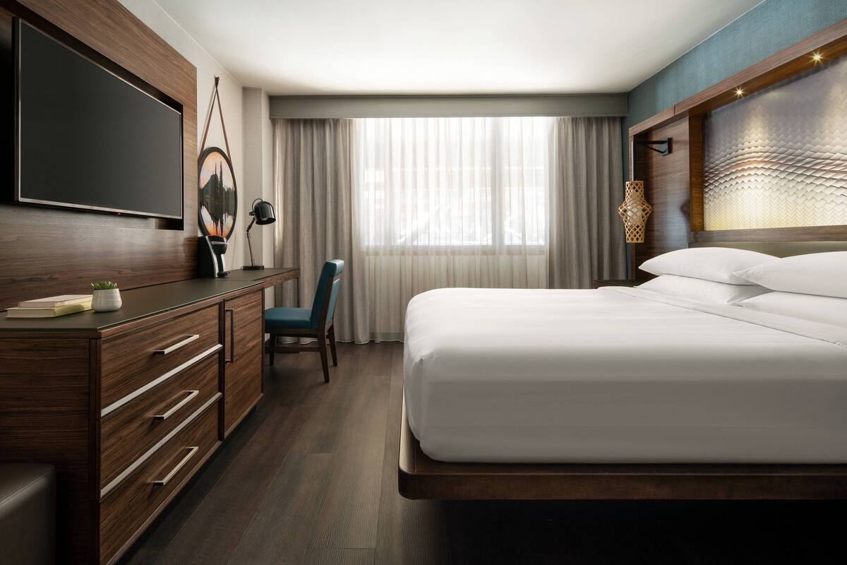 A hotel room with a large bed, a desk with drawers, a flat-screen TV mounted on the wall, and a large window with sheer curtains.