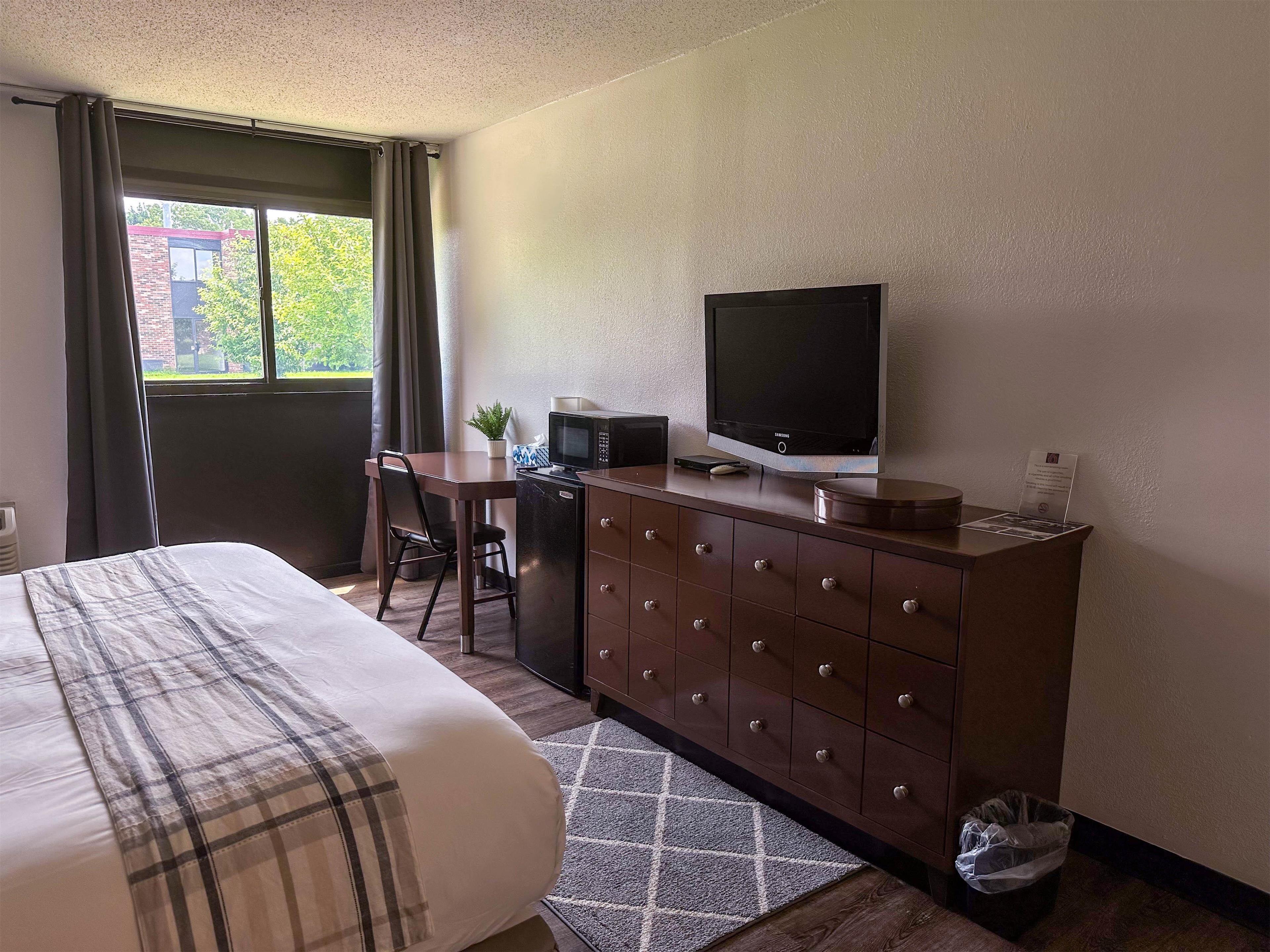 A hotel room with a bed, a desk with a chair, a television, a microwave, a dresser, and a window with curtains.