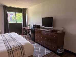 A hotel room with a bed, a desk with a chair, a television, a microwave, a dresser, and a window with curtains.
