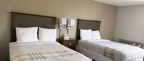Quarto de hotel com duas camas, cada uma com roupa de cama branca e manta xadrez. Há duas mesas de cabeceira com luminárias e uma caixa de lenços. O quarto tem uma paleta de cores neutras, com paredes bege e cabeceira de madeira.