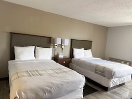 Habitación de hotel con dos camas, cada una con ropa blanca y una manta a cuadros. Hay dos mesitas de noche con lámparas y un dispensador de pañuelos. La habitación tiene una decoración en tonos neutros, con paredes beige y un cabecero de madera.