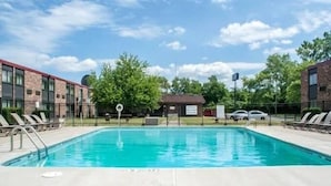 A rectangular swimming pool with a clear blue water surface, surrounded by a concrete deck. There are lounge chairs on the left side and a small building with a sign in the background. - Motel 6 Rochester, NY - Airport (Rochester)