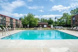 Una piscina rectangular con superficie de agua azul cristalina, rodeada por una solana de concreto. Hay sillas de descanso a la izquierda y un pequeño edificio con un letrero al fondo.