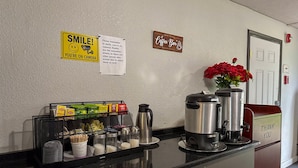 A coffee station with a sign that says "SMILE! YOU'RE ON CAMERA" and a note reminding guests to smile. There are coffee makers, a coffee pot, and a mug rack with snacks. - Motel 6 Rochester, NY - Airport (Rochester)