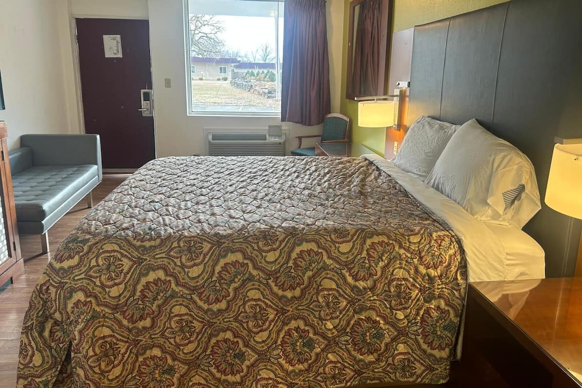 A hotel room with a bed, a chair, a desk, a window with curtains, and a door.