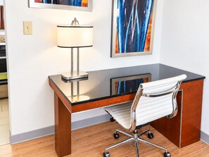 Uma mesa de escritório moderna com tampo de vidro, uma cadeira branca com estrutura cromada, uma luminária e arte abstrata na parede.