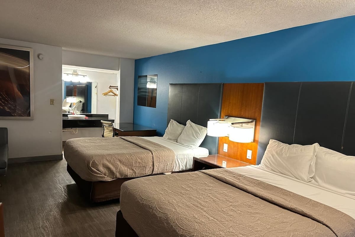 A hotel room with two beds, a desk, a mirror, and a nightstand with a lamp.