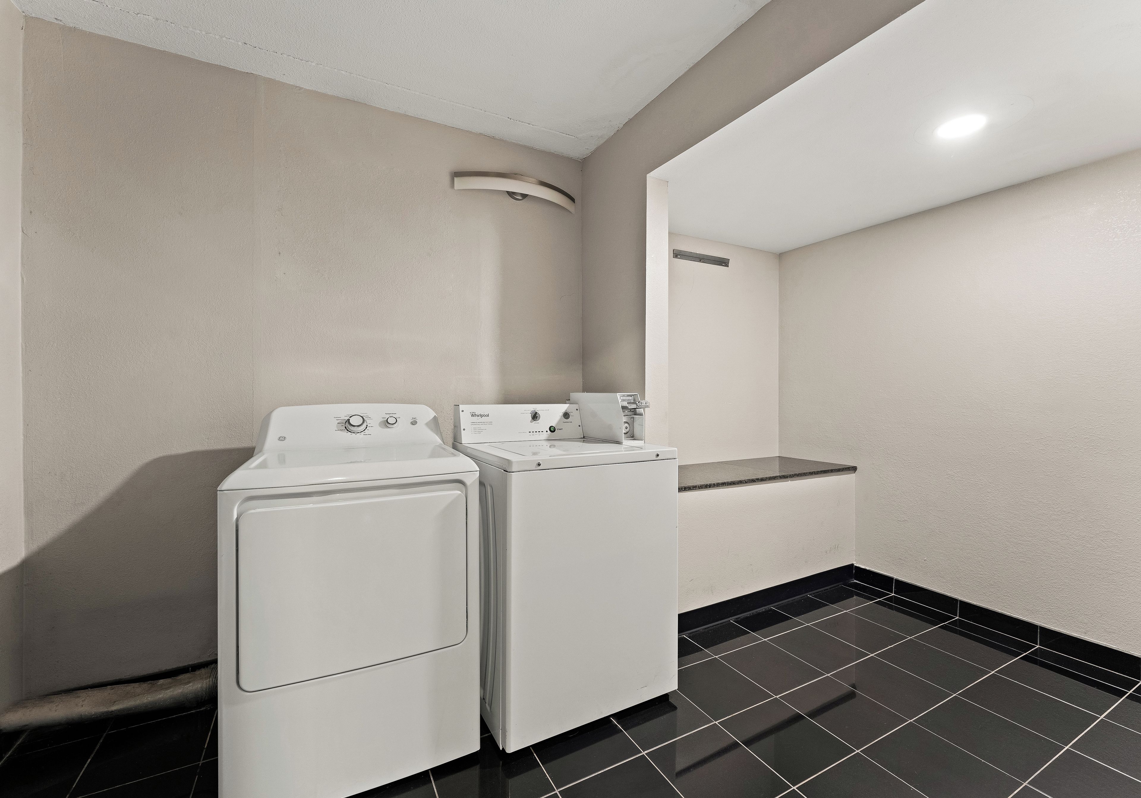 Un lavadero equipado con dos lavadoras y secadoras de carga frontal blancas, suelo de baldosas negras y techo blanco con una lámpara empotrada.