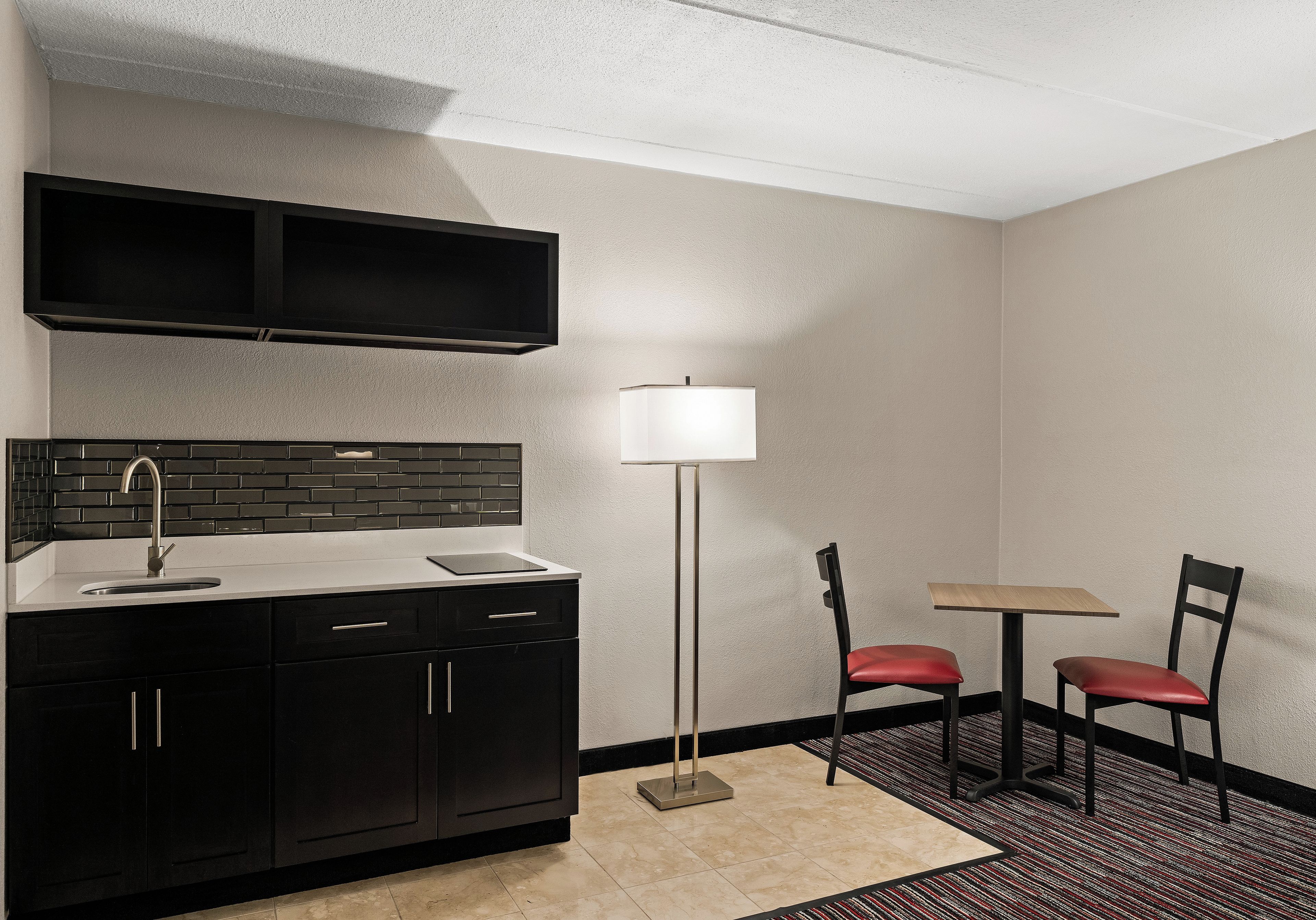 A compact kitchenette with a black countertop, a sink, and a backsplash. There is a small table with two chairs and a floor lamp.
