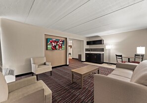 Habitación de hotel con televisión de pantalla plana en la pared, sofá, una silla, una mesita y una zona de kitchenette.
