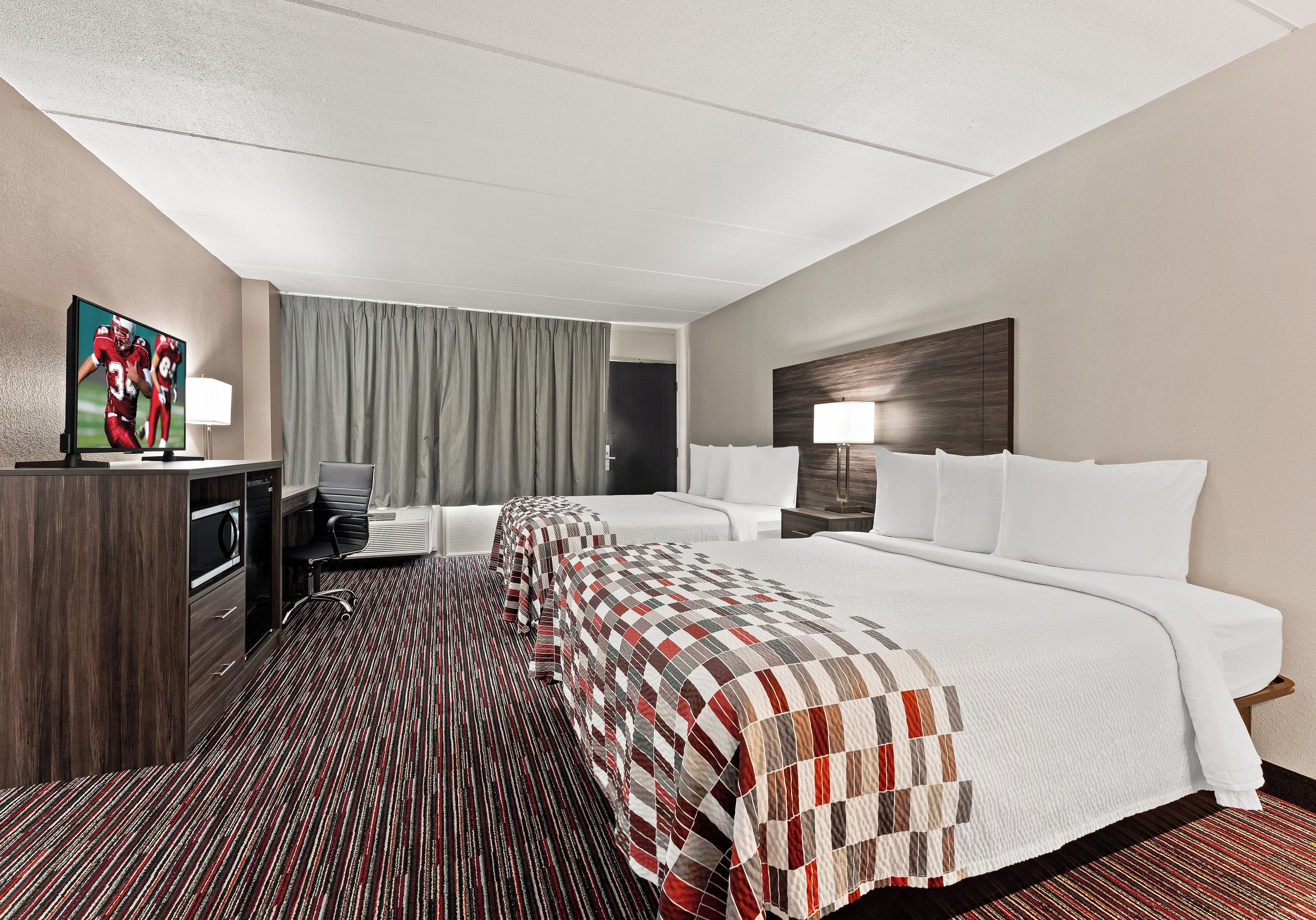 Ein Hotelzimmer mit einem großen Bett, einem Schreibtisch mit Stuhl, einem Fernseher und einem Fenster mit Vorhängen.