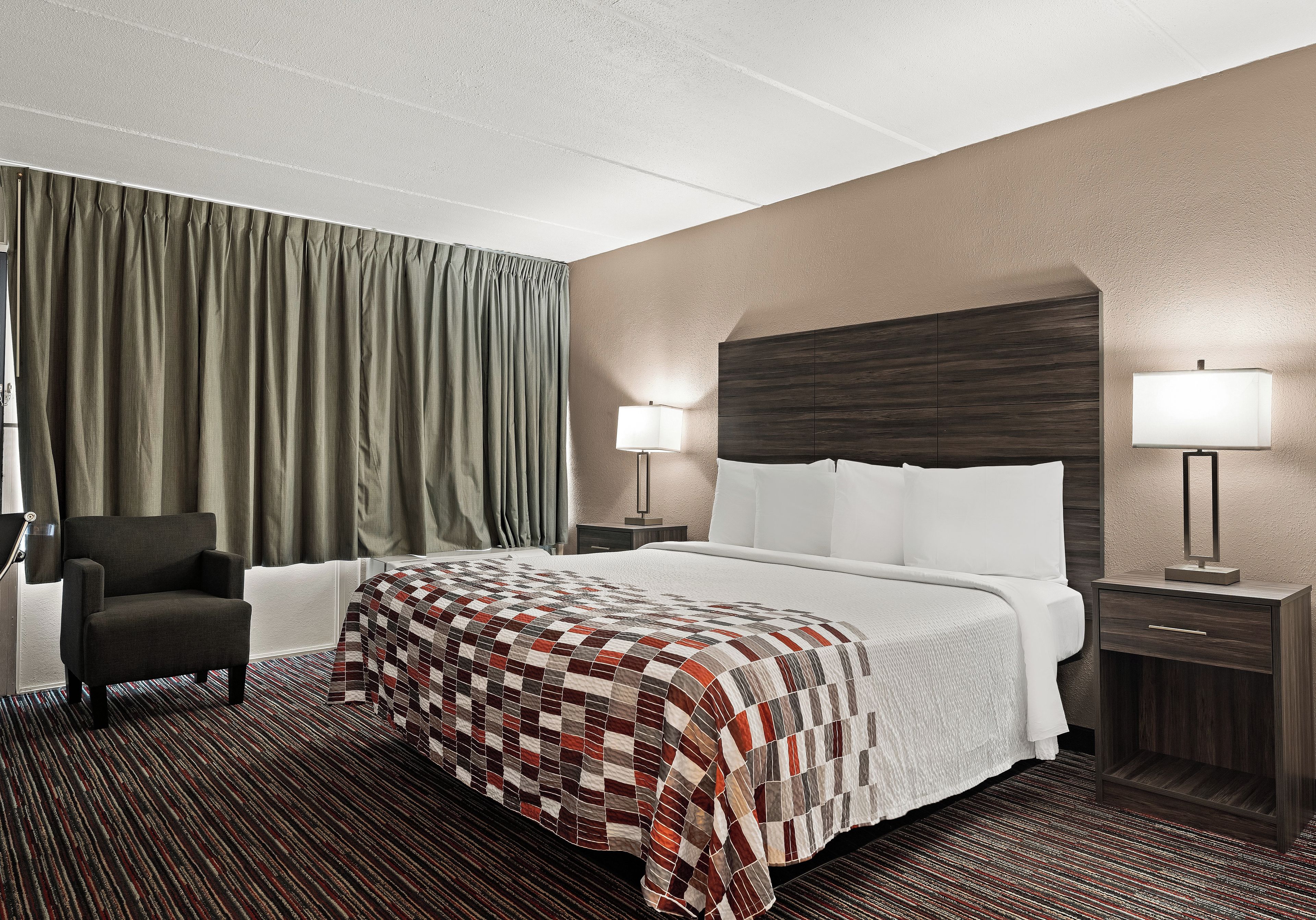 Une chambre d’hôtel avec un lit, une chaise, un bureau, une lampe et une fenêtre avec des rideaux.