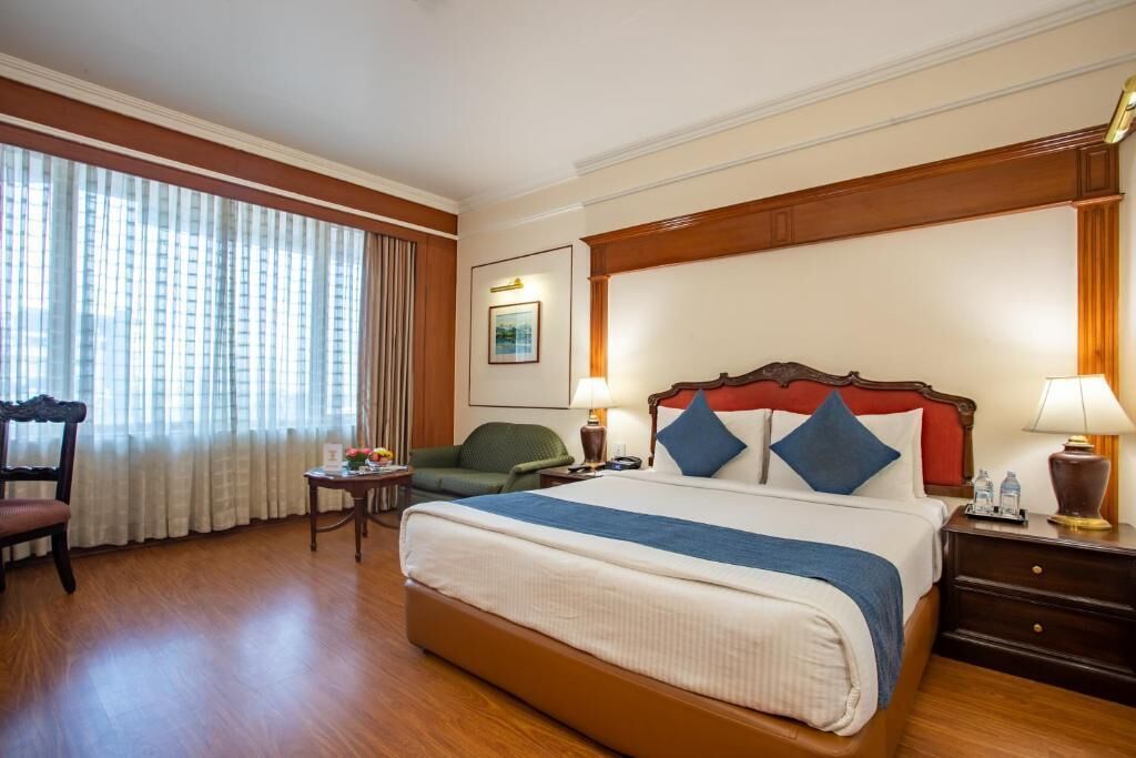 Quarto de hotel com uma cama grande, uma cadeira, uma escrivaninha e uma janela com cortinas.