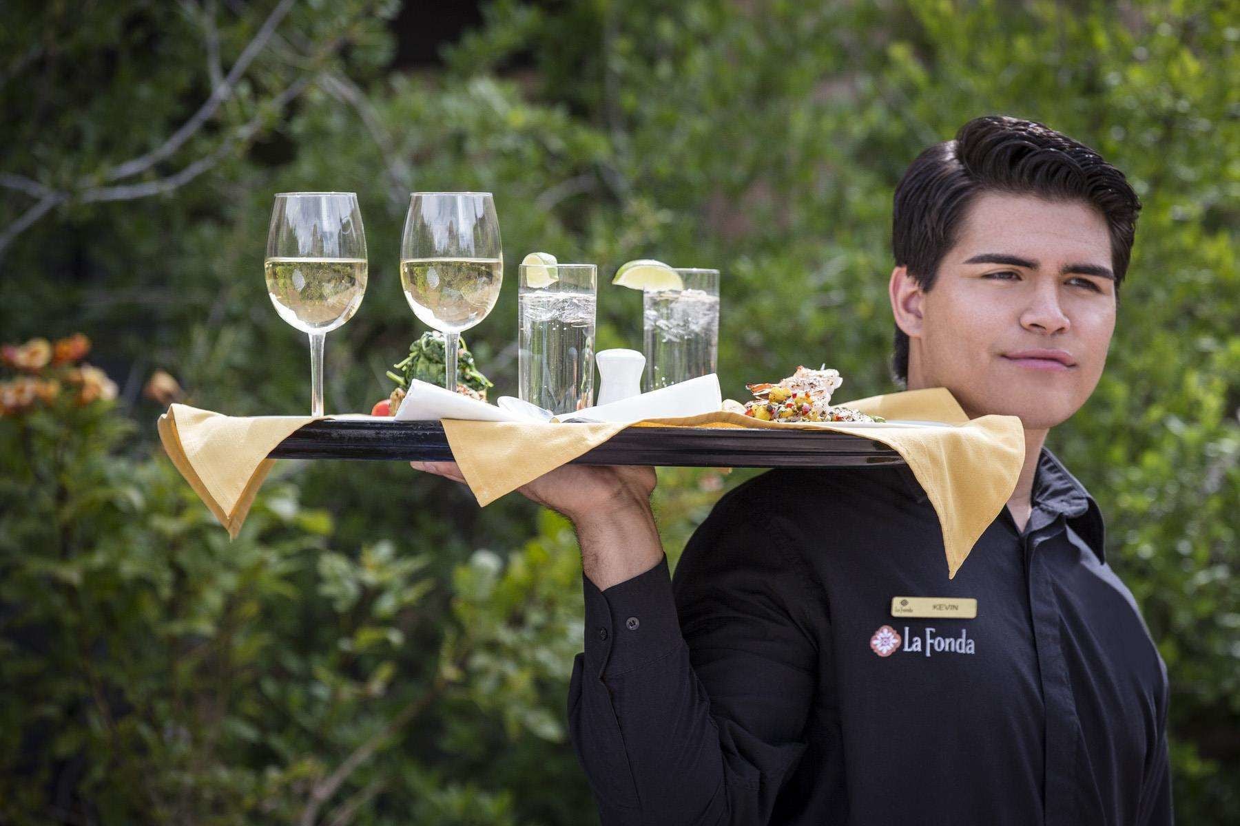 Une personne porte un plateau sur lequel se trouvent des verres de vin blanc, un citron et un plat de nourriture.