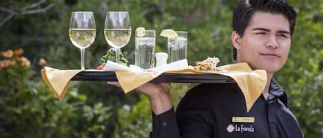 A person carrying a tray with glasses of white wine, a lemon, and a dish of food.