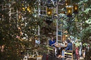 An indoor dining area with tables and chairs, surrounded by greenery and decorative lighting.