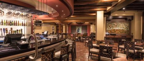 A bar area with a counter, stools, and a variety of bottles. There are tables and chairs arranged for dining.