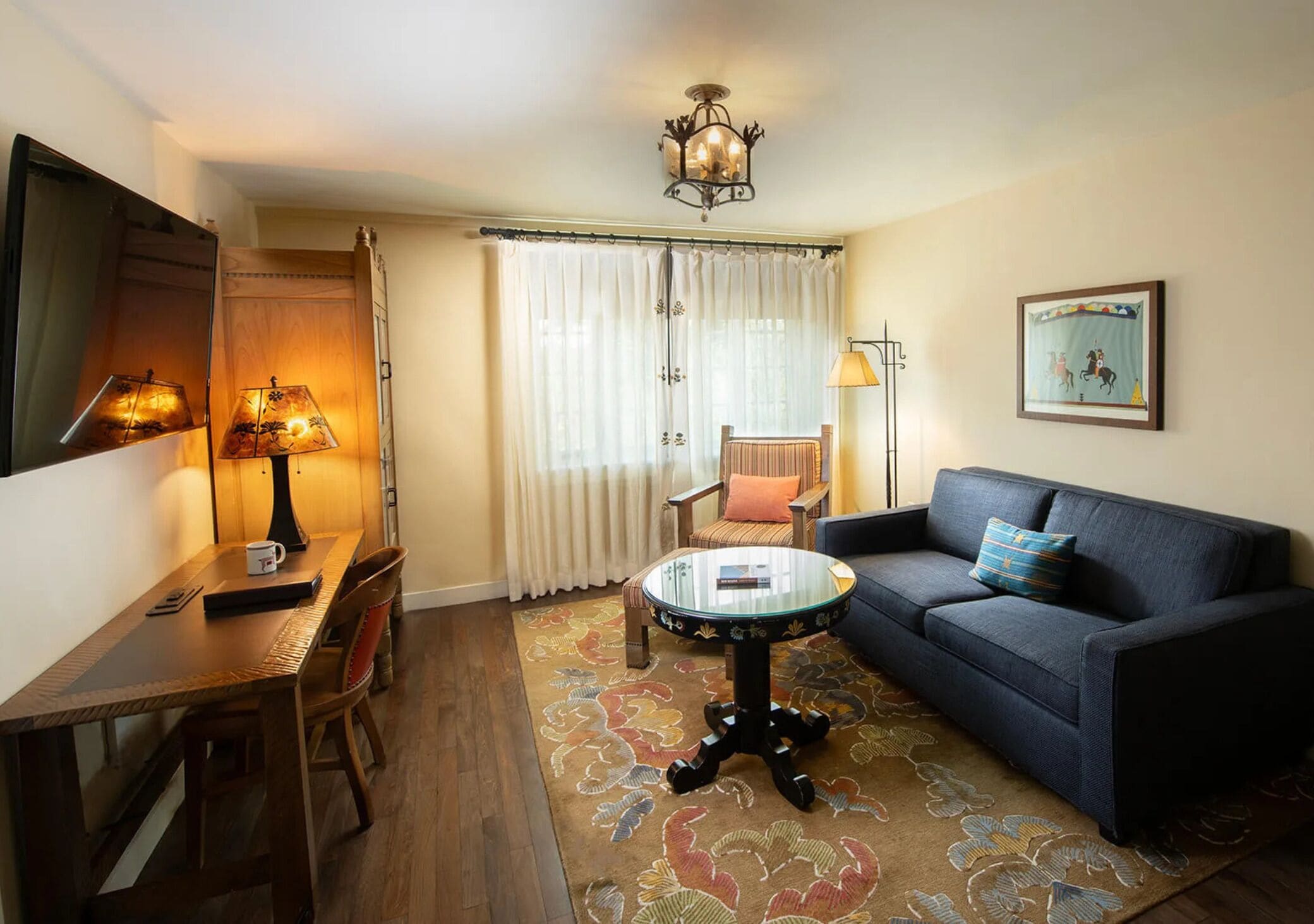 A cozy living room with a sofa, a small round coffee table, a dining table with chairs, a lamp, and a large window with curtains.